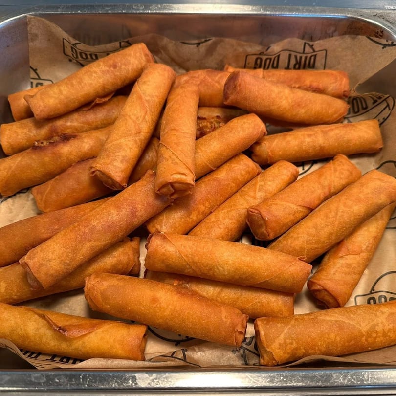 A metal tray filled with numerous golden-brown Filipino lumpia egg rolls on brown parchment paper.