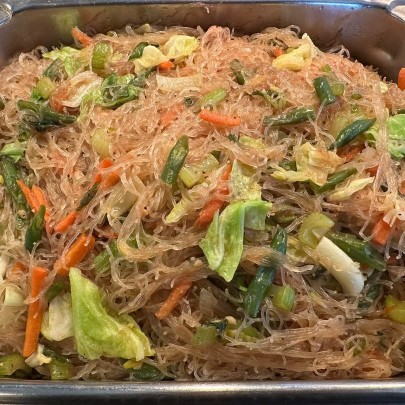 Close-up of Filipino pancit bihon noodles with carrots, cabbage, and green beans in a metal tray.